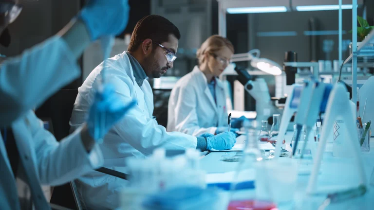 A scientist sits in a lab writing, with colleagues surrounding him.